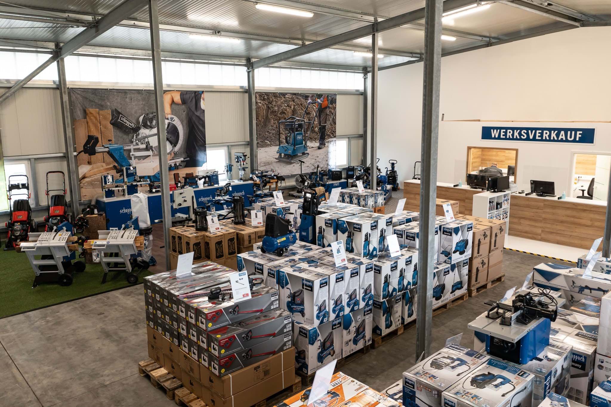 Scheppach-Werkverkauf mit verschiedenen Werkzeugen und Maschinen. Gezeigt werden unter anderem Schleifmaschinen, Bohrmaschinen und andere Elektrowerkzeuge. Die Produkte sind in Kartons verpackt und auf Paletten gestapelt.