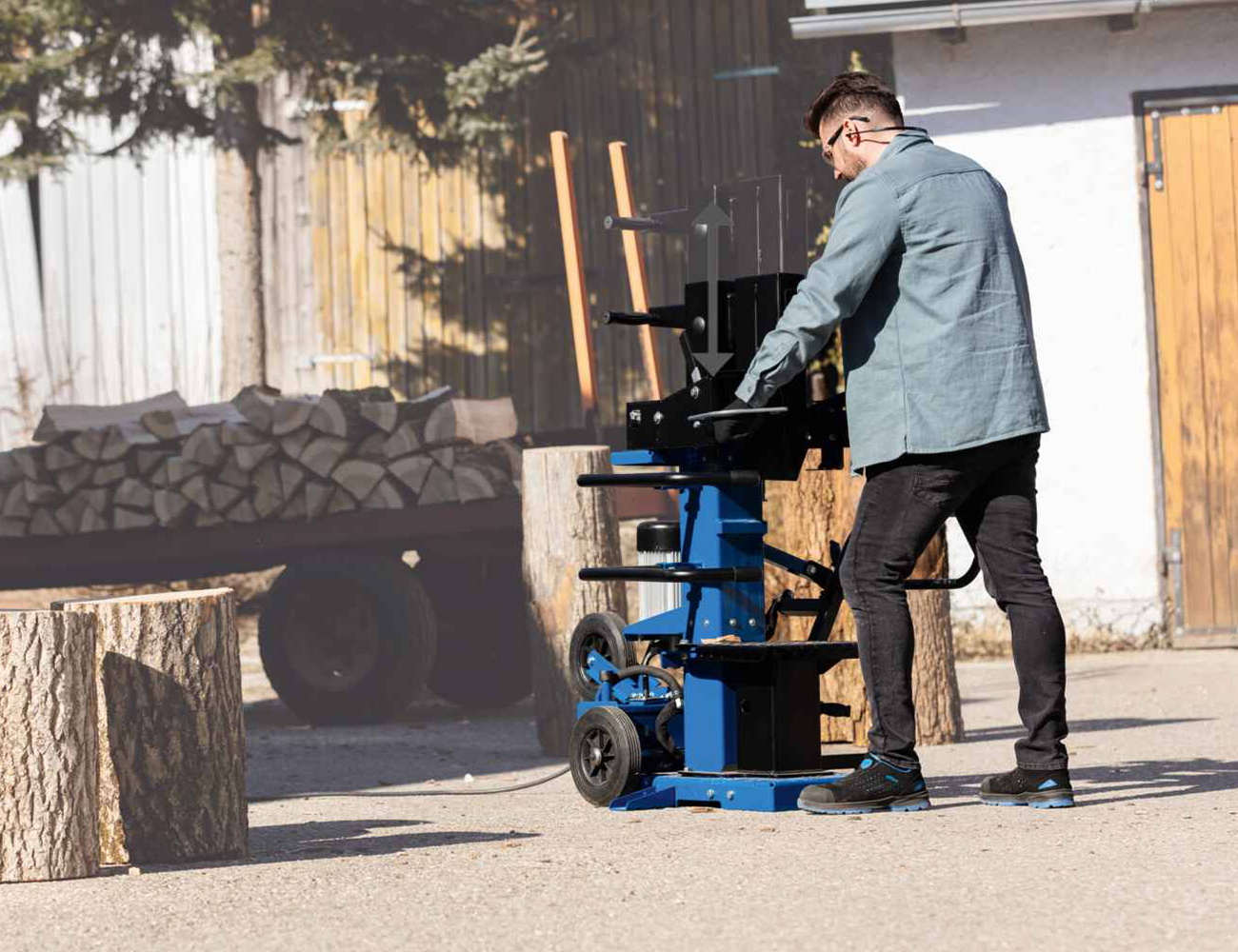 Ein Mann bedient einen Holzspalter Compact 15 von Scheppach. Der Holzspalter ist mobil und hat große Räder. Im Hintergrund sind Holzstapel zu sehen.