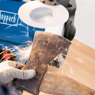 Ein Scheppach Doppelschleifer BG200AL wird zum Schleifen eines Holzstücks verwendet. Ein Handschuh ist sichtbar.