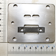 Ventilplatte mit Messskala im Hintergrund. Die Platte hat mehrere Löcher und Aussparungen. Die Platte wird von der Seite gezeigt.