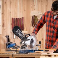 Ein Mann bedient eine Kapp- und Gehrungssäge HM80L von Scheppach in einer Werkstatt. Die Säge ist auf einem Holztisch montiert und schneidet durch ein Holzbrett. Im Hintergrund sind verschiedene Werkzeuge und Materialien zu sehen.