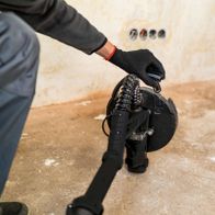 Ein Mann hält den Scheppach Wand- und Deckenschleifer DWS90X Woodster mit beiden Händen. Der Schleifer ist auf dem Boden positioniert.