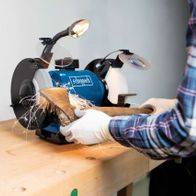 Scheppach Doppelschleifer BG200AL im Einsatz. Eine Hand hält ein Holzstück gegen die Schleifscheibe. Im Hintergrund weitere Werkzeuge.