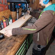 A man operates a Scheppach bench pillar drill DP16SL in a workshop. He wears safety goggles and ear protection. The machine features a laser for precise drilling.