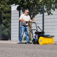 Ein Mann bedient eine Kehrmaschine SC2400p von Scheppach auf einem asphaltierten Weg. Die Maschine hat einen gelben Besen und einen Auffangbehälter.