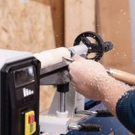 Scheppach Drechselmaschine DM1100T im Einsatz. Ein Werkstück wird mit einem Holzstab bearbeitet. Holzspäne fliegen durch die Luft. Im Hintergrund sind Holzplatten zu sehen.