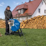 Ein Mann bedient die Wippsäge HS510 von Scheppach im Garten. Im Hintergrund sind Holzstapel und ein Haus zu sehen.