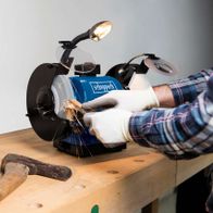 Ein Benutzer bearbeitet Metall mit dem Scheppach Doppelschleifer BG200AL. Werkzeuge wie Hammer und Meißel liegen auf der Werkbank. Der Schleifer ist in Betrieb und erzeugt Funken.