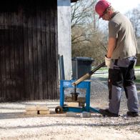 Ein Arbeiter bedient einen Steinknacker HSC130 von Scheppach. Der Mann trägt eine rote Schutzhelm und Arbeitskleidung. Im Hintergrund ist ein Gewässer und eine kleine Hütte zu sehen.