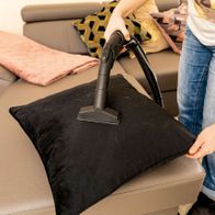 Eine Person reinigt ein Sofa mit dem Scheppach SprayVac20. Der schwarze Aufsatz liegt auf einem Kissen. Der Sauger ist an einem Schlauch befestigt.