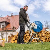 Ein Mann bedient eine Wippsäge HS410 von Scheppach im Garten. Im Hintergrund sind Holzstapel zu sehen.