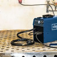 Scheppach Multischweißgerät WSE4000-Multi auf perforiertem Metalltisch. Kabel und Schlauch liegen auf dem Tisch. Im Hintergrund sind Gasflaschen und Schläuche zu sehen.