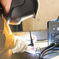 Ein Arbeiter verwendet das 3in1 Kombi-Schweißgerät WSE4000-CUT von Scheppach. Der Arbeiter trägt eine Schweißerhaube und Handschuhe und hält einen Schweißbrenner in der Hand. Das Gerät zeigt eine Temperatur von 147 an.