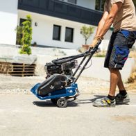 Ein Mann bedient eine Scheppach Rüttelplatte HP1300S auf einer Betonfläche. Die Maschine hat einen schwarzen Motor und ein blaues Gehäuse. Im Hintergrund sind ein Gebäude und Paletten zu sehen.