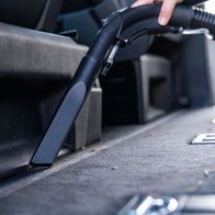 Ein Scheppach SprayVac20-Sauger, der in den Kofferraum eines Autos gehalten wird. Der Sauger hat einen schwarzen Griff und einen grauen Behälter. Der Sauger wird von einer Hand gehalten, die den Griff festhält.
