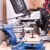 Kappzugsäge HM254 von Scheppach im Einsatz. Ein Benutzer führt ein Holzstück durch die Säge. Die Säge ist auf einem blauen Tisch montiert und schneidet durch das Holz. Der Benutzer trägt eine rote und schwarze Kariertjacke.