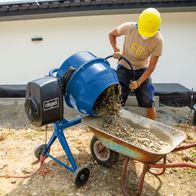 Ein Arbeiter mit gelbem Helm bedient einen Betonmischer von Scheppach. Der Mischer füllt eine Schubkarre mit Beton. Der Mischer hat ein blaues Gehäuse und einen schwarzen Motor mit dem Scheppach-Logo. Die Szene spielt auf einer Baustelle.