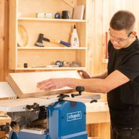 Ein Mann bedient die Abricht- & Dickenhobelmaschine HMS1070 von Scheppach. Er trägt Schutzbrille und bearbeitet ein Holzbrett. Im Hintergrund sind Werkzeuge und Holzregale zu sehen.