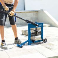 Ein Mann bedient einen Steinknacker HSC130 von Scheppach. Er trägt Schutzhandschuhe und -brille und drückt den Hebel des Geräts nach unten. Der Steinknacker steht auf einer Betonplatte.