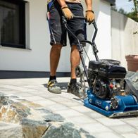 Ein Mann bedient eine Scheppach Rüttelplatte HP1300S auf einem gepflasterten Boden. Die Maschine hat einen schwarzen Motor und eine blaue Basis. Im Hintergrund sind ein Blumentopf und eine Hauswand zu sehen.