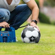 Ein Mann sitzt auf einem Rasen und verwendet einen Scheppach-Kompressor, um einen Fußball aufzublasen. Der Kompressor ist blau und hat ein digitales Druckdisplay.