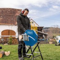 Ein Mann bedient eine Wippsäge HS410 von Scheppach im Freien. Die Säge ist auf einem Ständer montiert und wird zum Schneiden von Holzstämmen verwendet. Im Hintergrund sind ein Schuppen und weitere Werkzeuge zu sehen.