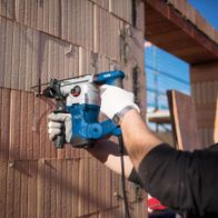 Scheppach DH1300 Plus hammer drill in use on a wall. A worker holds the tool with both hands and wears protective gloves.