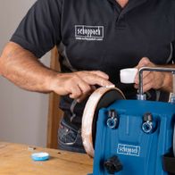 Ein Mann bedient eine Scheppach-Nass-Schleifmaschine. Er hält einen Schleifstein und führt ihn an der Maschine aus. Die Maschine ist blau und trägt den Namen 'Scheppach'.
