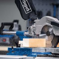 Kappzugsäge HM110MP von Scheppach im Einsatz. Ein Benutzer schneidet ein Holzbrett. Die Säge ist auf einem blauen und schwarzen Tisch montiert.