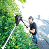 Ein Mann schneidet mit einer Elektro Heckenschere TPX710 von Scheppach einen grünen Strauch. Er trägt Schutzbrille und Kopfhörer.