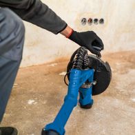 Wand-Deckenschleifer DS920X von Scheppach im Einsatz. Person hält das Gerät mit Handschuhen und führt es über den Boden. Der Schleifer hat einen blauen Griff und eine schwarze Schleifscheibe.
