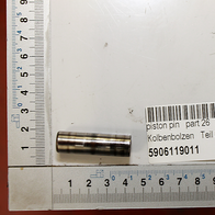Ein Kolbenbolzen mit der Bezeichnung 'Kolbenbolzen Teil 26' und der Artikelnummer 5906119011. Der Bolzen liegt neben einem Maßstab und einem Beschriftungslabel. Der Kolbenbolzen ist von der Seite zu sehen.