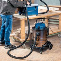 Der Scheppach Nass- und Trockensauger ASP50-ES mit 50L, HEPA-Filter, Steckdose und Einschaltautomatik. Der Sauger ist mit einem Holzfäller verbunden, um Sägespäne zu entfernen.