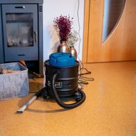 Aschesauger AVC20 Scheppach mit Zubehör und Holzbehälter neben einem Kamin. Der Aschesauger ist mit einem feuerfesten Schlauch ausgestattet und steht auf dem Boden. Der Aschesauger ist von der Seite zu sehen.