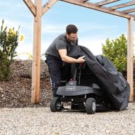 Ein Mann deckt einen Scheppach Aufsitzrasenmäher mit einer schwarzen Abdeckung ab. Der Rasenmäher steht unter einem Holzpavillon.
