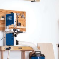 Scheppach DC100 dust extractor with vacuum and hose, mounted on a workbench with wooden shelves and tools in the background.