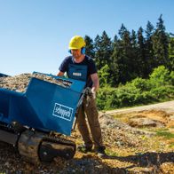Kettendumper DP5000 Scheppach mit 500 kg Traglast. Ein Arbeiter in Schutzausrüstung steht neben dem beladenen Dumper auf einem Baustellengelände. Im Hintergrund sind Bäume und eine unbefestigte Straße zu sehen.