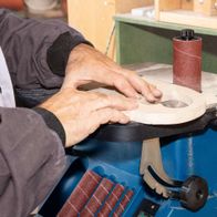 Ein Mann bedient einen Scheppach OSM600 Oszillierender Kanten-, Band- und Spindelschleifer. Er hält ein Werkstück gegen die rotierende Schleifscheibe.