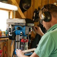 A man operates a Scheppach bench pillar drill DP19Vario. He wears safety goggles and ear protection. The machine has a transparent housing and a red handle.