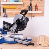 Kappzugsäge HM254 von Scheppach auf einer Werkbank mit verschiedenen Werkzeugen im Hintergrund. Die Säge hat einen schwarzen Griff und eine graue Basis. Die Werkzeuge sind an einer Holzwand befestigt.