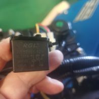 Ein Hand hält ein Relais für Kontrollpanel RTT7101-12V / JD1914 / 40A von Scheppach. Das Relais ist schwarz und hat Beschriftungen mit technischen Details. Im Hintergrund ist ein blaues Kontrollpanel zu sehen.