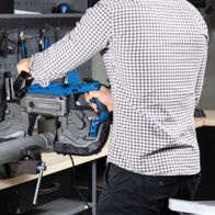 Metallbandsäge MBS1200xt von Scheppach, seitliche Ansicht. Ein Mann hält die Bandsäge fest, während sie auf einem Tisch montiert ist. Im Hintergrund sind Werkzeuge an der Wand befestigt.