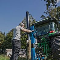 Ein Mann bedient einen Holzspalter HL3000GM von Scheppach. Der Holzspalter ist an einen Traktor angeschlossen und wird auf einer Wiese verwendet.