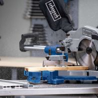 Kappzugsäge HM110MP von Scheppach im Einsatz. Ein Benutzer führt ein Holzstück durch die Säge. Die Säge ist auf einem blauen Tisch montiert und schneidet präzise.