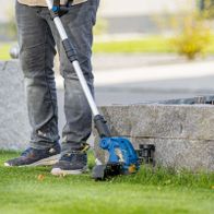 Person mit Gehstock nutzt Scheppach-Akku-Rasentrimmer für Gartenpflege, mit Höhenverstellungsmarkierung und 3 Meter Fadenlänge.