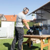 Ein Mann bedient einen liegenden Holzspalter HL460 von Scheppach. Der Holzspalter ist auf einem Holztisch platziert und hat einen Käfig für Holzscheite. Im Hintergrund befindet sich ein Gebäude mit einem Kirchturm.
