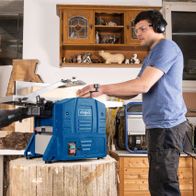 Ein Mann bedient eine Scheppach Abricht- und Dickenhobel HMS860 in einer Werkstatt. Die Maschine hat einen blauen Gehäuseteil und einen weißen Gehäuseteil. Im Hintergrund sind Holzstücke und ein Regal mit Werkzeugen zu sehen.