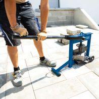 Steinknacker HSC130 von Scheppach im Einsatz. Ein Arbeiter bedient die Maschine auf einer Terrasse. Im Hintergrund sind Pflastersteine und Werkzeuge zu sehen.