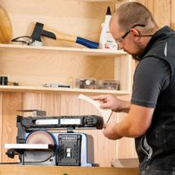 Ein Mann bedient einen Scheppach Band- und Tellerschleifer BTS900. Im Hintergrund sind Werkzeuge und Materialien zu sehen.