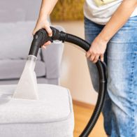 Ein Mensch reinigt mit dem Scheppach SprayVac20 einen hellgrauen Sessel. Der Sauger hat einen transparenten Aufsatz und einen schwarzen Schlauch.